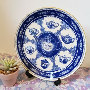 May include: A blue and white ceramic plate with a gold rim features a teapot design with the text "The Teapot Collection". The plate is decorated with eight smaller teapots in a circular pattern.