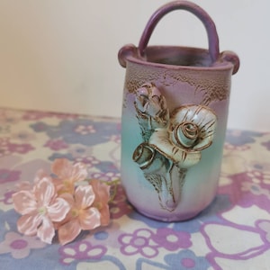 Pode incluir: Um pequeno vaso de cerâmica com alça, pintado em um degradê de azul claro para roxo. O vaso tem um design de rosa 3D na frente.