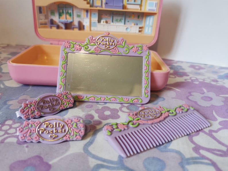 Puede incluir: Un set de juego Polly Pocket morado con un espejo, dos pinzas para el pelo y un peine. El espejo tiene un dise&ntilde;o floral y el texto "Polly Pocket" en &eacute;l. Las pinzas para el pelo y el peine tambi&eacute;n tienen el texto "Polly Pocket" en ellos.