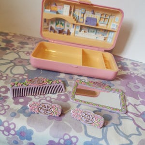 Puede incluir: Un set de juego Polly Pocket rosa con una casa en el interior, un peine, un espejo y dos pinzas para el pelo. La casa tiene una cocina, una sala de estar y un dormitorio. Las pinzas para el pelo tienen el logotipo de Polly Pocket.