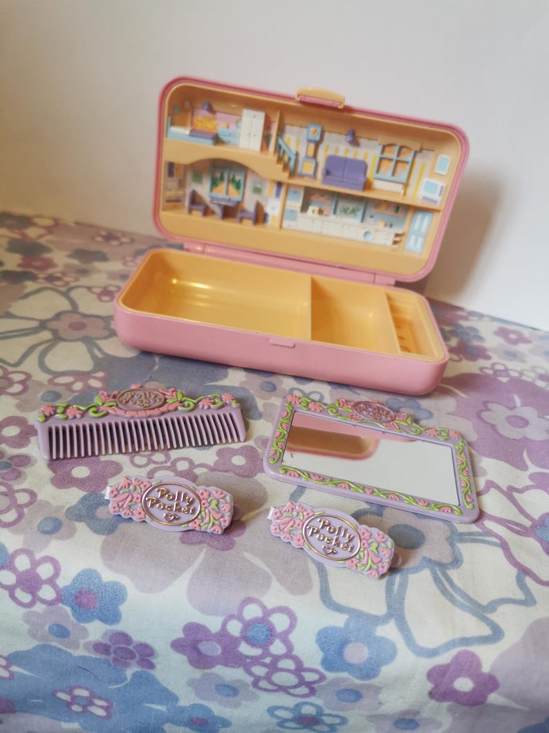 Puede incluir: Un set de juego Polly Pocket rosa con una casa en el interior, un peine, un espejo y dos pinzas para el pelo. La casa tiene una cocina, una sala de estar y un dormitorio. Las pinzas para el pelo tienen el logotipo de Polly Pocket.