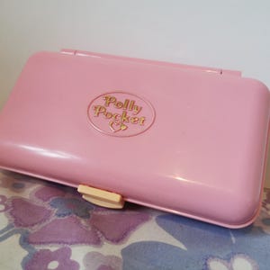 Puede incluir: Una caja rosa Polly Pocket con un logotipo dorado que dice "Polly Pocket" con un s&iacute;mbolo de coraz&oacute;n.