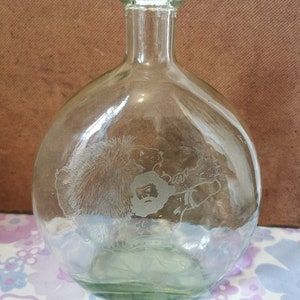 Puede incluir: Una botella de vidrio transparente con cuerpo redondo y cuello estrecho. La botella está grabada con un diseño de un hombre con barba y un oso.