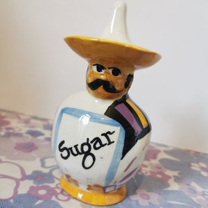 May include: A ceramic sugar dispenser shaped like a man wearing a yellow sombrero. The man has a black moustache and a purple and black patterned shirt. The dispenser says "Sugar" on the front.