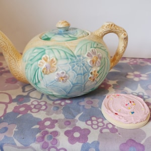 Puede incluir: Una tetera de cerámica con un diseño floral en tonos verdes, azules y rosas. La tetera tiene un patrón de telaraña y un asa con forma de rama. Una galleta glaseada rosa con chispas de colores está sobre un mantel floral.