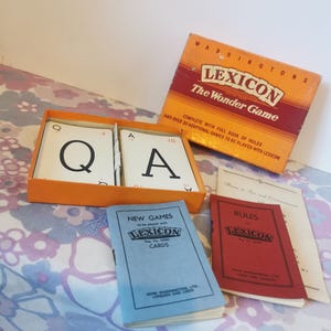 May include: An orange box set of the Lexicon word game, with a red and orange box lid that reads "LEXICON The Wonder Game". The set includes letter cards, rule books, and a blue booklet titled "NEW GAMES".