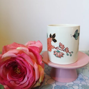 Puede incluir: Taza de cerámica blanca con un diseño floral con flores rosas y naranjas, hojas verdes y una mariposa azul. La taza está sobre un pedestal rosa.