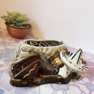 Może przedstawiać: Ceramiczna figurka przedstawiająca śpiącą postać z sombrero. Postać jest brązowo-biała, z sombrero obok. Figurka ma wbudowaną doniczkę z metalowymi cewkami. Mała roślina sukulent w miedzianej doniczce znajduje się w tle.