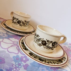 May include: Two cream-colored teacups and saucers with a brown floral design. The cups have a handle and sit on matching saucers. The saucers have a brown rim and a floral pattern. The set is on a floral patterned surface.
