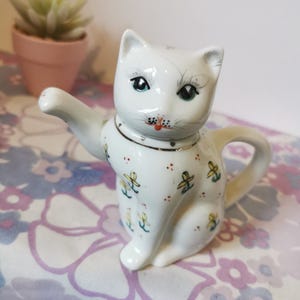 May include: A white ceramic cat-shaped teapot with a raised paw. The teapot has a gold rim and is decorated with small yellow and blue floral designs. The cat's face has blue eyes, a red nose, and black whiskers. A small succulent plant in a pink pot is in the background.