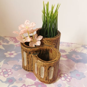 May include: A brown ceramic planter with three separate compartments, two of which are filled with artificial plants. The planter has a unique heart shape and is decorated with white stripes.