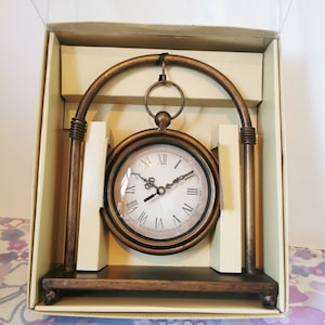 Peut inclure: Une horloge dorée avec des chiffres romains sur un support décoratif. L'horloge est dans une boîte avec un couvercle en plastique transparent.