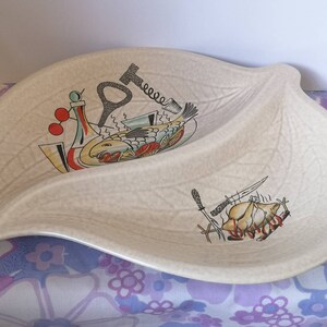 Może przedstawiać: Biała ceramiczna tacka do serwowania w kształcie liścia z dwoma sekcjami. Tacka jest ozdobiona czarno-czerwoną ilustracją ryby i kurczaka, oba z nożami i innymi narzędziami kuchennymi.