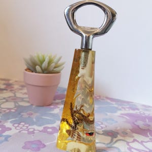 Puede incluir: Un abridor de botellas de plástico transparente amarillo con una parte superior de metal plateado. El abridor de botellas tiene una concha y un cangrejo dentro del plástico.