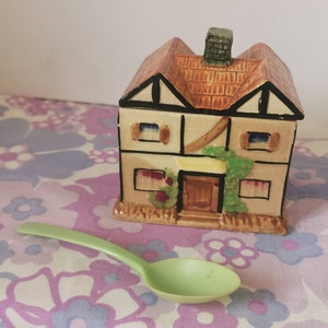 Puede incluir: Un azucarero de cerámica con forma de casa con un techo marrón, paredes blancas y hiedra verde. El bol tiene una pequeña cuchara de plástico verde a su lado.