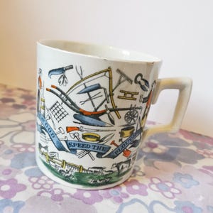 Puede incluir: Taza de cerámica blanca con una ilustración colorida de herramientas agrícolas y el texto "God Speed the Plough".