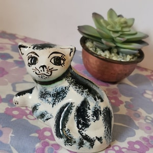 May include: A ceramic figurine of a black and white cat with painted eyes and a brown mouth. The cat is sitting on a floral patterned fabric.