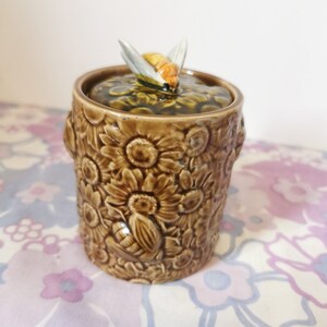 May include: A brown ceramic honey pot with a bee on top. The pot is decorated with a floral pattern.