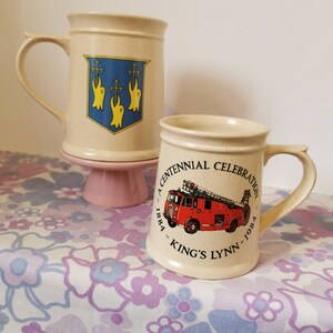 May include: Two ceramic mugs. The first mug is beige with a blue and yellow crest featuring three crosses and three yellow birds. The second mug is beige with a red fire truck and the text "A Centennial Celebration 1884 - King's Lynn - 1984".