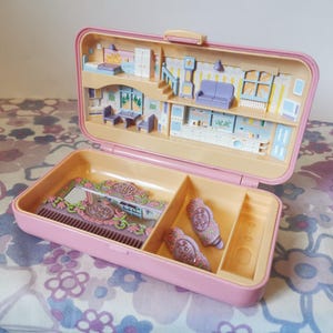 Puede incluir: Una casa de mu&ntilde;ecas de pl&aacute;stico rosa con un espejo, un peine y pinzas para el pelo. La casa tiene un dormitorio, una cocina y una sala de estar. La casa est&aacute; abierta y el interior es visible.