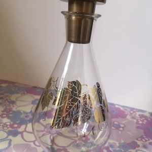 May include: A clear glass decanter with a gold leaf design. The decanter has a round body and a tall, thin neck. The top of the decanter has a gold-coloured stopper.