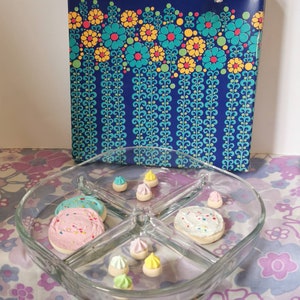 May include: A clear glass divided serving dish with four sections. The dish is filled with small cookies and candies. The cookies are pink, blue, and white with sprinkles. The candies are yellow, green, and white. The dish is sitting on a floral tablecloth.