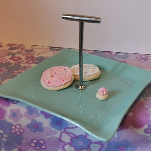 May include: A teal square cake stand with a silver metal stand and three cookies. Two cookies have pink and white frosting with sprinkles. One cookie has white frosting with sprinkles. A small pink frosted cookie is on the stand.