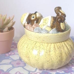 May include: A ceramic cookie jar shaped like a yellow sack with a brown and white animal peeking out of the top. The animal is wearing a red hat and has a brown tail.