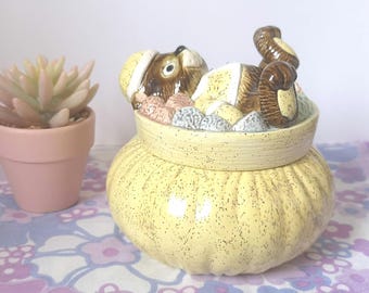May include: A ceramic cookie jar shaped like a yellow sack with a brown and white animal peeking out of the top. The animal is wearing a red hat and has a brown tail.