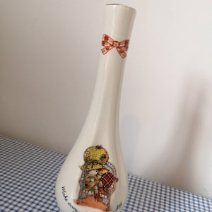 May include: White ceramic vase with a red and yellow bow at the top. The vase has a floral design with a girl sitting in a chair and the text "make every day a happy day".