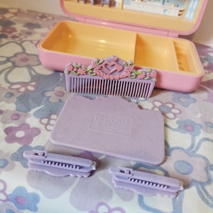Puede incluir: Un peine de pl&aacute;stico morado con un dise&ntilde;o floral y un coraz&oacute;n en el centro. Hay dos pinzas para el pelo de pl&aacute;stico morado y una bandeja de pl&aacute;stico morado con las palabras "Bluebird Toys" impresas en ella.