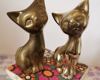 May include: Two gold-colored metal cat figurines. The cats are sitting on a colorful floral patterned surface.