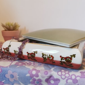 Puede incluir: Una caja de porcelana blanca para alhajas con una tapa verde y un diseño floral. La caja tiene una banda rosa con detalles dorados y flores rojas. Un collar de cuentas moradas descansa sobre la caja.