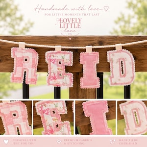 May include: A fabric banner spelling the name REID in pink and white letters. Each letter features a different pattern, including gingham, floral, and solid pink fabric, with scalloped edges. The letters are suspended from a white rope.
