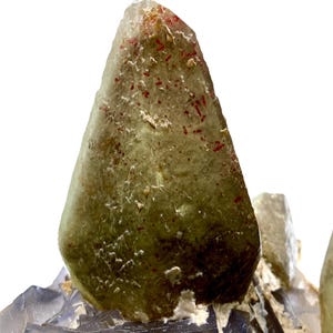 Puede incluir: Primer plano de un cristal transl&uacute;cido de color verde claro con inclusiones rojas. El cristal tiene forma piramidal puntiaguda y se asienta sobre un grupo de formaciones minerales grises y azules. La apariencia general es natural y geol&oacute;gica.