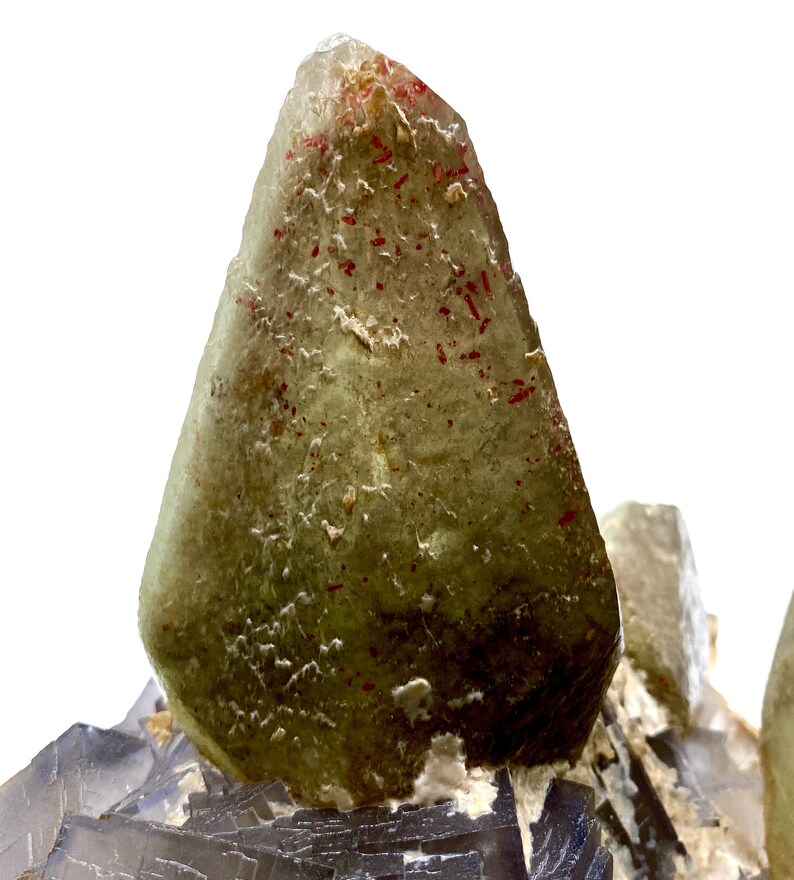 Puede incluir: Primer plano de un cristal transl&uacute;cido de color verde claro con inclusiones rojas. El cristal tiene forma piramidal puntiaguda y se asienta sobre un grupo de formaciones minerales grises y azules. La apariencia general es natural y geol&oacute;gica.