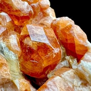 Puede incluir: Primer plano de un grupo de cristales de calcita naranja. Los cristales tienen una apariencia translúcida y vidriosa con bordes afilados y un color vibrante y cálido. El fondo es negro, resaltando los cristales.