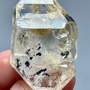 Puede incluir: Un cristal de cuarzo transparente con inclusiones negras. El cristal está doblemente terminado y tiene una forma puntiaguda.