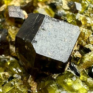 May include: Close-up of a dark, faceted mineral specimen with a cubic shape. The dark crystal is surrounded by smaller, yellow-green crystals. The lighting highlights the mineral's texture and the surrounding crystals.