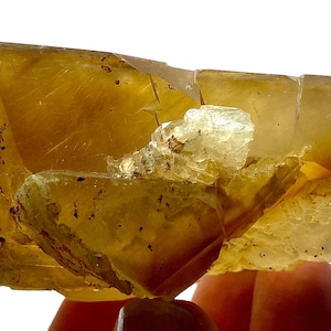 Op de afbeelding: Een doorschijnend, amberkleurig mineraal specimen met een kristallijne structuur. Het mineraal heeft een gelaagde uitstraling met een lichtere, kristallijne formatie bovenop. Het specimen wordt vastgehouden door de vingers van een persoon.