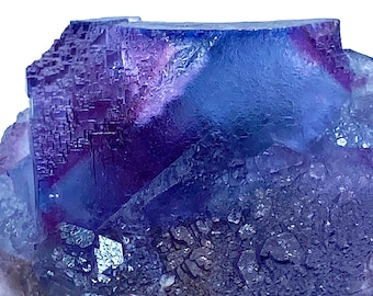 May include: A close-up of a fluorite crystal with a layered appearance. The crystal features a gradient of colors, including deep purple, blue, and hints of red. The surface has a textured, crystalline structure, with a dark, rough base.