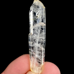 Puede incluir: Un cristal claro y alargado con una punta y un tono ligeramente amarillo. El cristal tiene estructuras internas y facetas visibles, sobre un fondo negro. La gema mide aproximadamente 7.6 cm de largo.