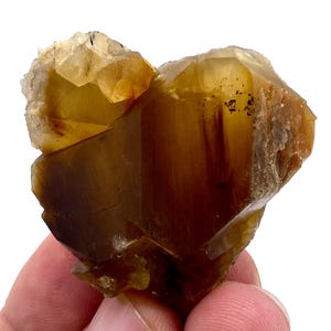 May include: A raw, natural mineral specimen with a warm, amber color. The crystal formation has a layered appearance with a mix of translucent and opaque sections. The edges are rough and uneven, showcasing the stone's natural state.