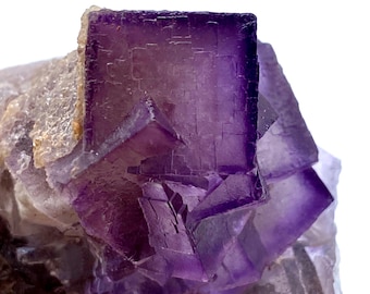 May include: Close-up of a purple fluorite crystal cluster. The crystal features a large, square-shaped formation with a deep purple hue, surrounded by lighter, translucent crystal structures. The edges of the crystal are sharply defined.