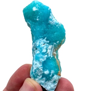 Puede incluir: Un vibrante espécimen mineral de turquesa en bruto con una superficie rugosa y texturizada. La piedra presenta áreas de azul brillante y blanco, con toques de marrón. La formación natural se sostiene en una mano sobre un fondo blanco.
