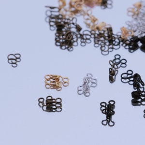 Könnte beinhalten: Eine Auswahl an Metall-Haken- und Ösenverschlüssen in Gold, Silber und Schwarz. Die Haken und Ösen sind auf einer weißen Oberfläche verstreut, einige befinden sich in einem rosa Behälter.