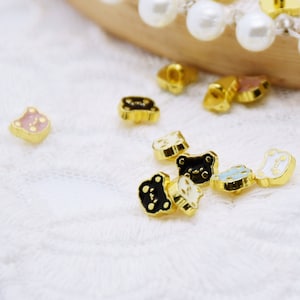 May include: Small, gold-colored bear-shaped charms with enamel details in pink, black, and blue. The charms are scattered on a white lace surface, some near a wooden circle trimmed with pearls. These decorative charms are ideal for jewelry making.