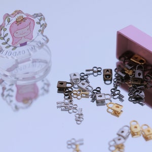 Puede incluir: Una caja rosa con una variedad de pequeñas cerraduras y llaves doradas, plateadas y negras que se derraman. La caja está sobre una superficie reflectante.