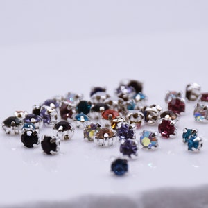 May include: Assortment of colorful rhinestones with silver settings. The rhinestones are scattered on a white surface.