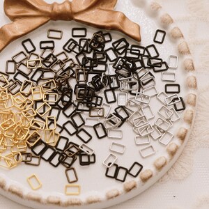 May include: A white ceramic dish with a gold bow on top, filled with a variety of small, rectangular metal jump rings in gold, silver, and black.